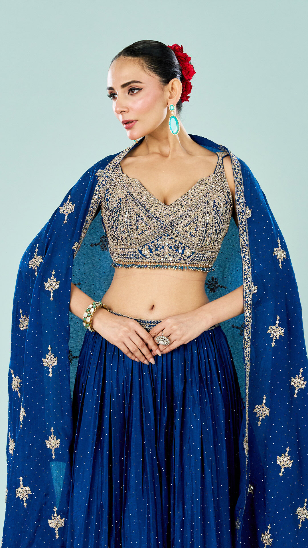 Bride wearing an embellished navy blue lehenga with a heavily embroidered sleeveless blouse and matching dupatta, styled with a red floral bun and turquoise earrings against a pastel blue backdrop.